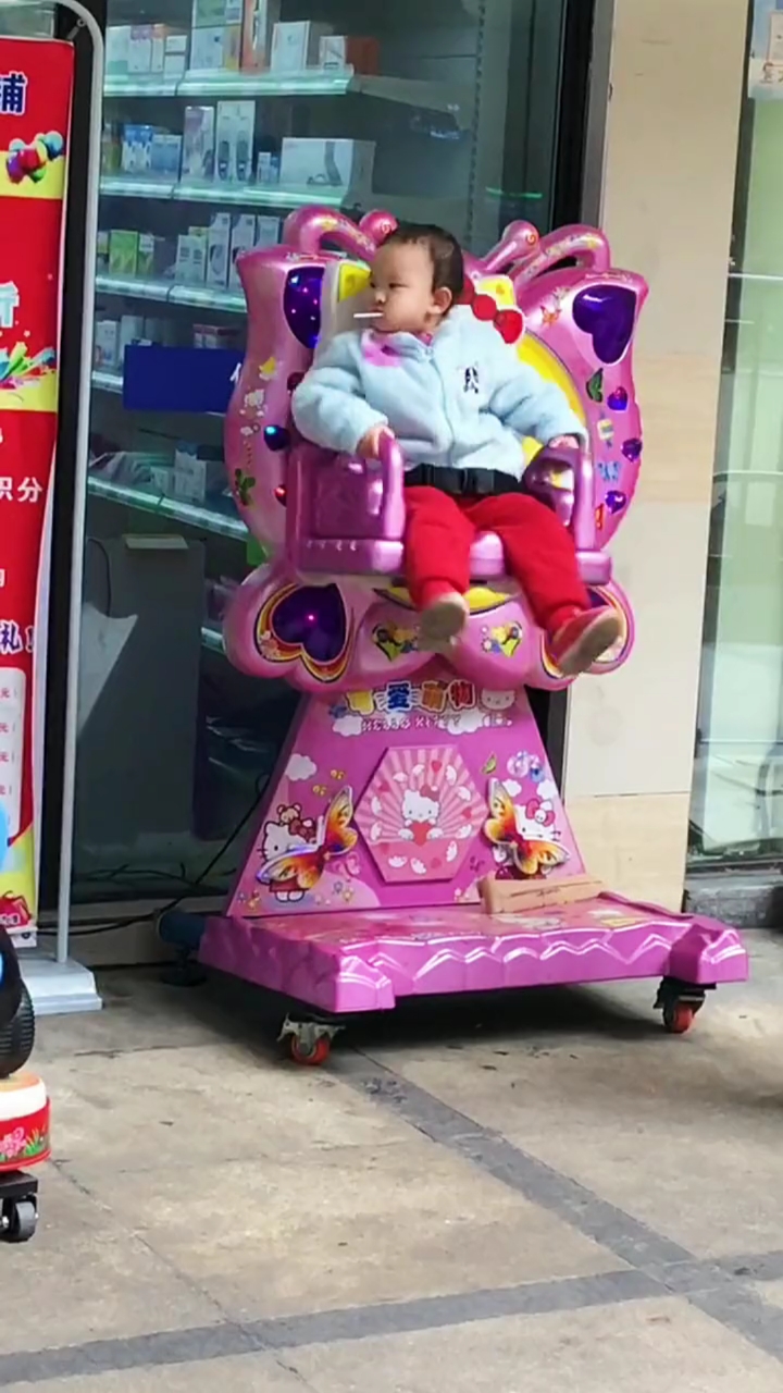 孩子奶奶在一旁和别人聊天,孩子一个人坐摇摇车玩,嘴里含着糖,看起来